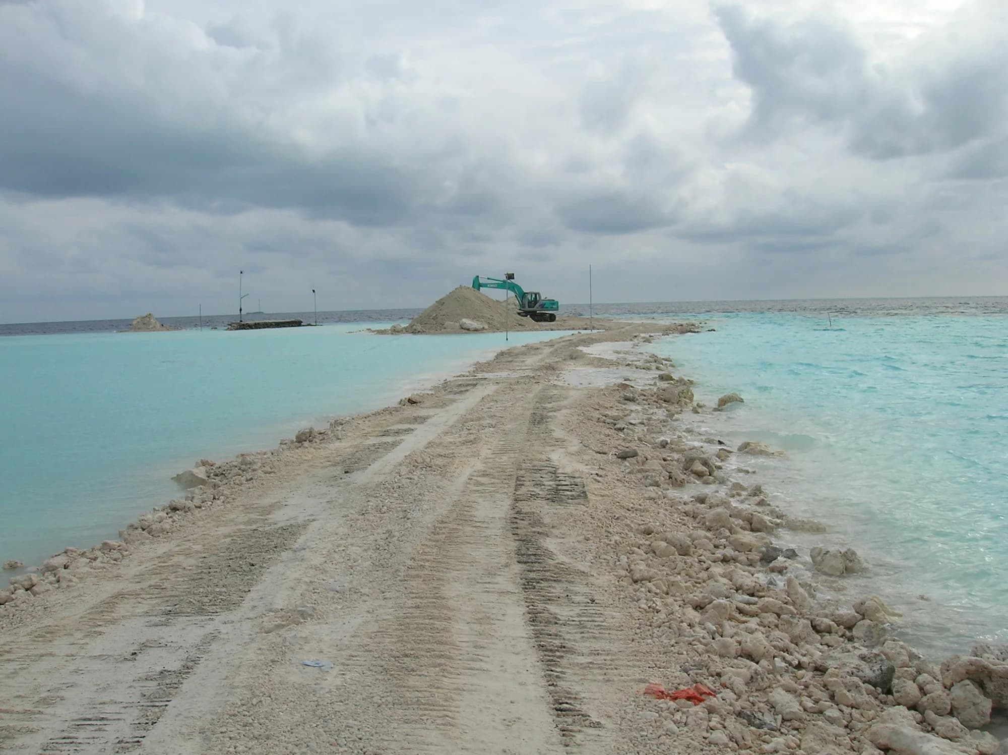 A sand haul road pushed out across a turquoise lagoon toward a working excavator